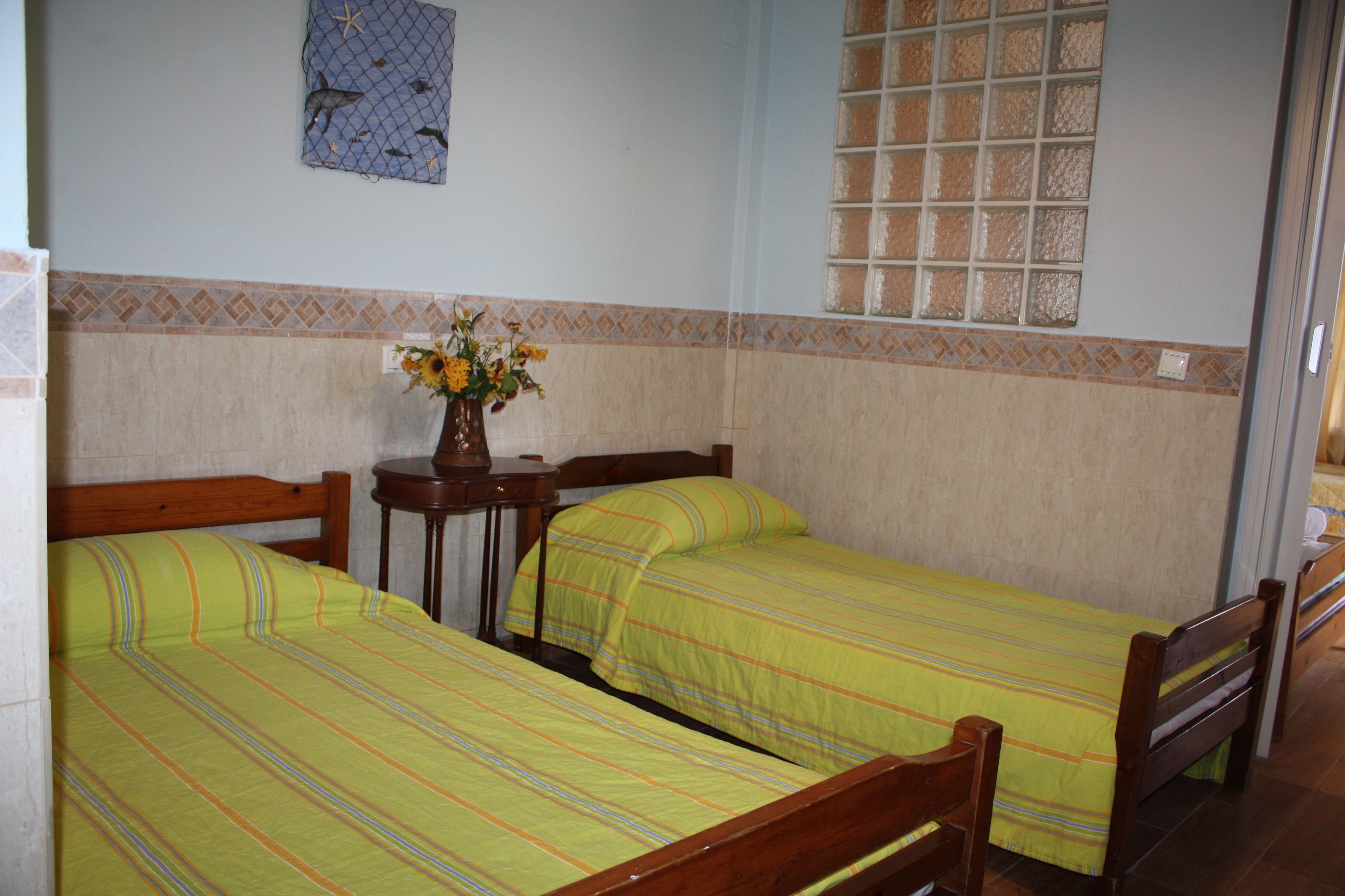 A room with two single beds, a small table with a vase of flowers, and a window with a grid pattern.