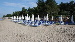 A beach with rows of blue and white beach chairs and umbrellas, a sandy shore, and trees in the background. - Villa Bambola (Nea Propontida)