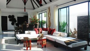 Una sala de estar moderna con un sofá, una mesa de centro de vidrio y un área de comedor con mesa y sillas. Hay un ventanal grande que proporciona luz natural y vistas a la vegetación exterior.