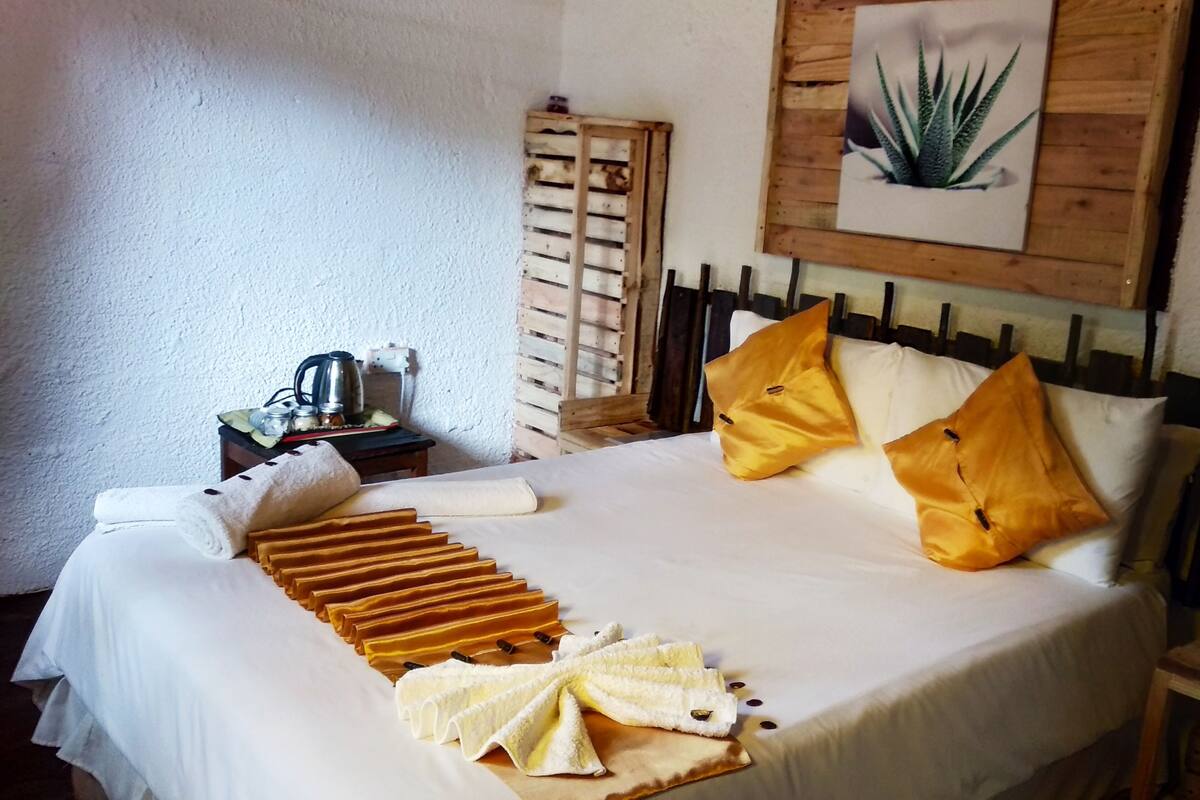 A neatly made bed with pillows and a folded towel, a wooden headboard, a framed plant on the wall, and a small table with a kettle and jars.