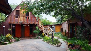 Una casa de madera rústica con un techo empinado, una escalera de madera que lleva a un porche, y un jardín con diversas plantas y un perro pequeño.
