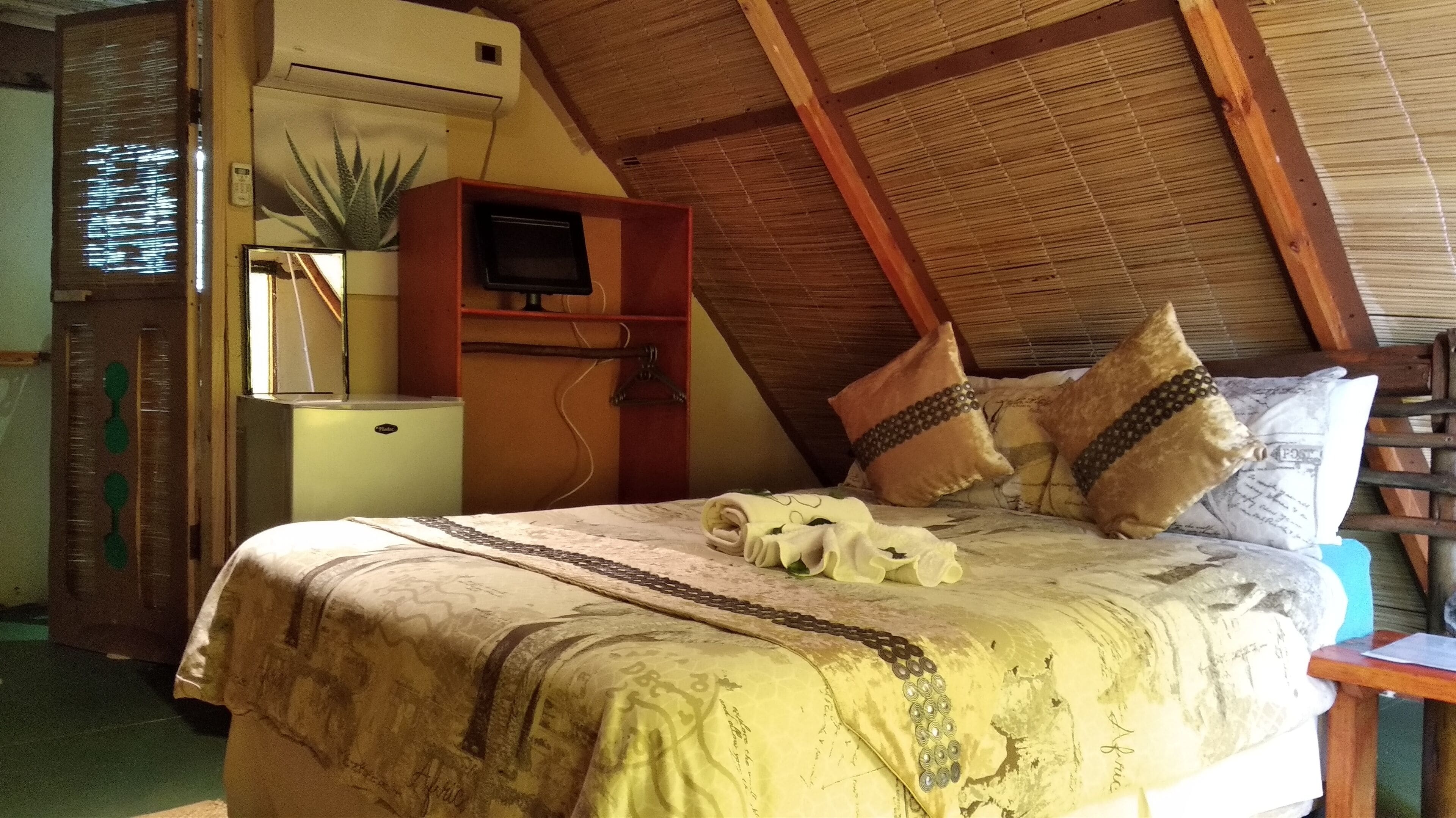 A bedroom with a bed, a wooden ceiling, a small desk, a plant, and a wall-mounted air conditioner.
