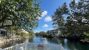 Eine Holzterrasse mit weißen Geländern, die auf einen ruhigen Fluss mit Bäumen und Gebäuden blickt.