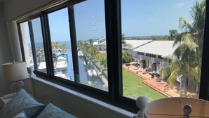Ein Zimmer mit großem Fenster mit Blick auf eine Marina und einen Ferienort. Es gibt ein Bett mit gemusterten Kissen und eine Lampe auf einem Nachttisch.