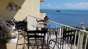 A balcony with a glass table and chairs, overlooking a clear blue sea with a distant island. - SPECTACULAR OCEANFRONT VIEWS , FREE WIFI, HD SMART TV'S, SUMMER SPECIAL RATES (Tavernier)
