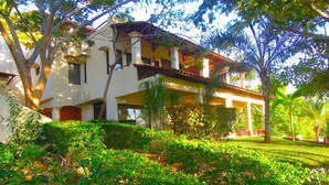 Um edifício de dois andares com varanda, cercado por vegetação exuberante e árvores.