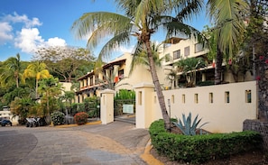 A gated entrance to a residential area with multiple buildings, palm trees, and a paved driveway. - OCEAN VIEW COMFORTABLE CONDO BY THE OWNER, 6 POOLS. COCO, COSTA RICA. (Playas Del Coco)