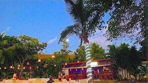 Uma área de estar ao ar livre com mesas e cadeiras brancas, um palco com artistas e um edifício com telhado vermelho.