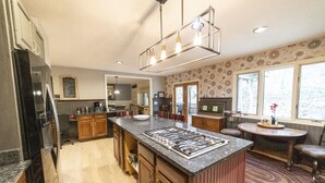 A modern kitchen with a central island, wooden cabinets, a stainless steel refrigerator, and a dining area with a round table and chairs. - We weathered Helene beautifully!      Welcome to your new, lovely mountain home! (Asheville)