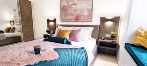 Ein modernes Schlafzimmer mit einem großen Bett, flauschigen Kissen, einem Nachttisch mit Lampe und einem an der Wand hängenden abstrakten Gemälde.