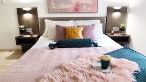 Ein ordentlich eingerichtetes Schlafzimmer mit einem großen Bett, zwei Nachttischen mit Lampen, einem an der Wand hängenden abstrakten Gemälde und einem flauschigen rosa Teppich.