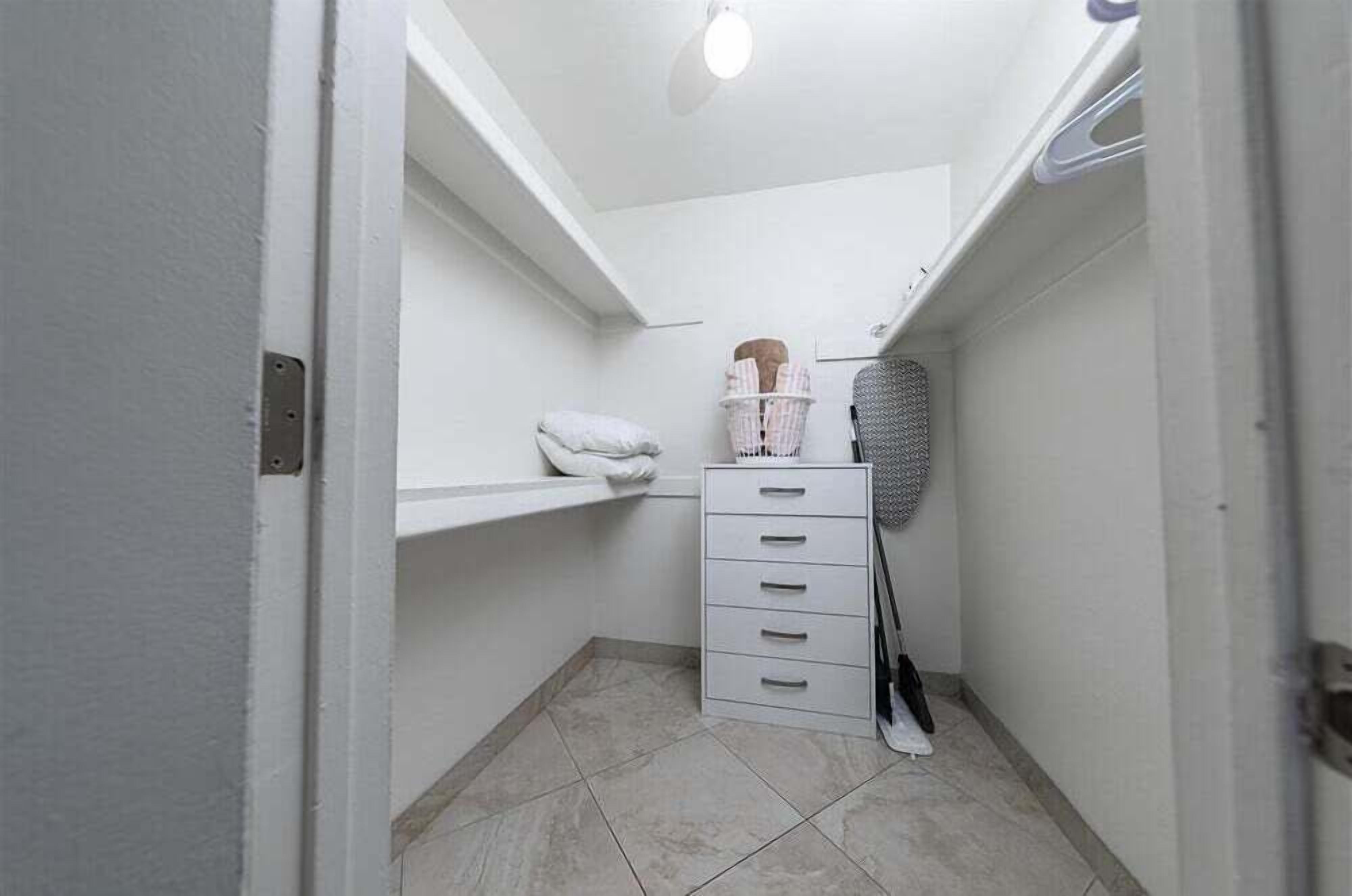 A narrow hallway with a white wall, a ceiling light, a white shelf with a pillow, a basket, a black chair, and a white drawer unit.