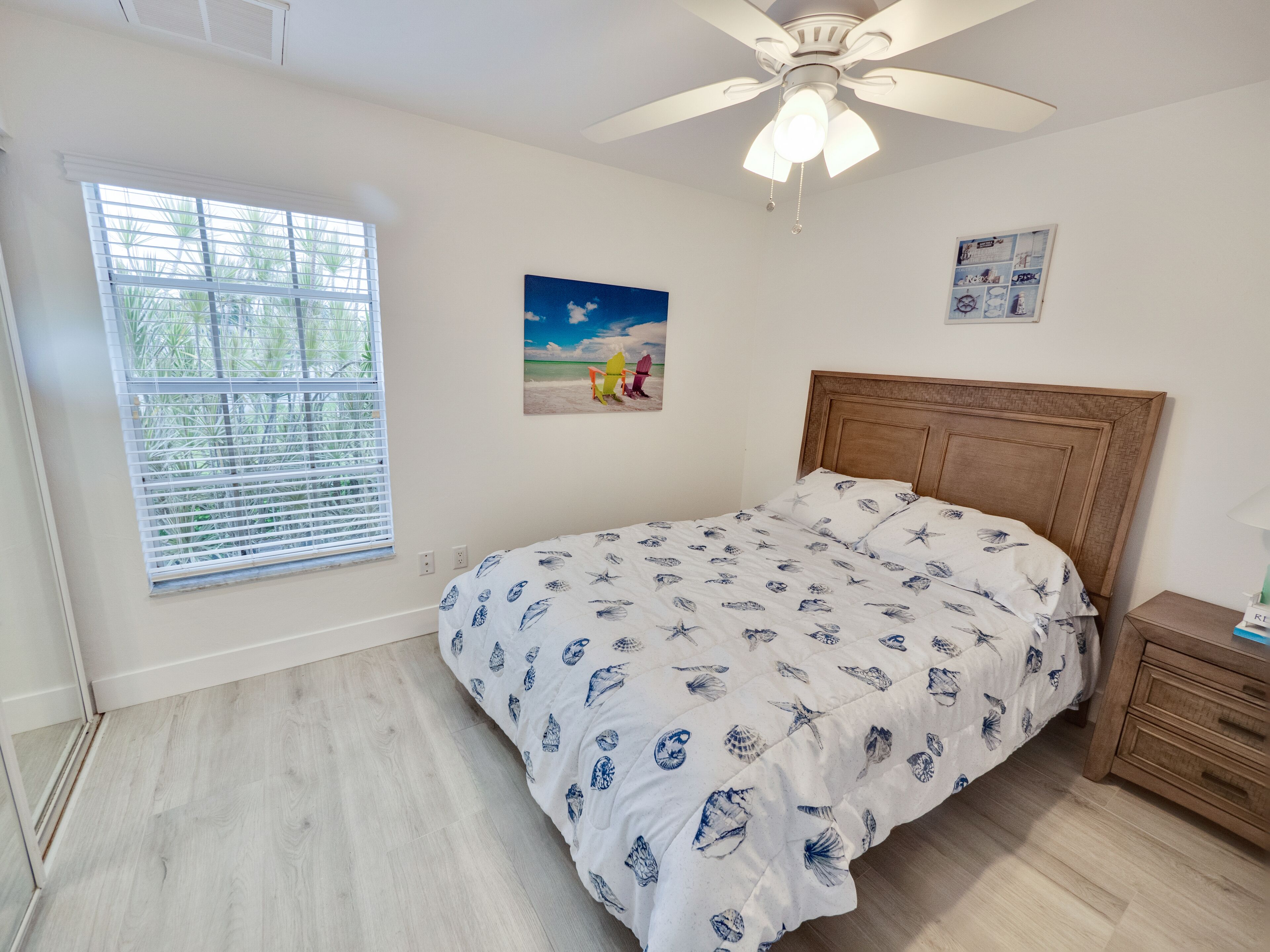A bedroom with a bed, a ceiling fan, a window with blinds, and a small bedside table.