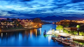 A riverside town with a calm river, boats docked along the shore, and buildings with lights on.