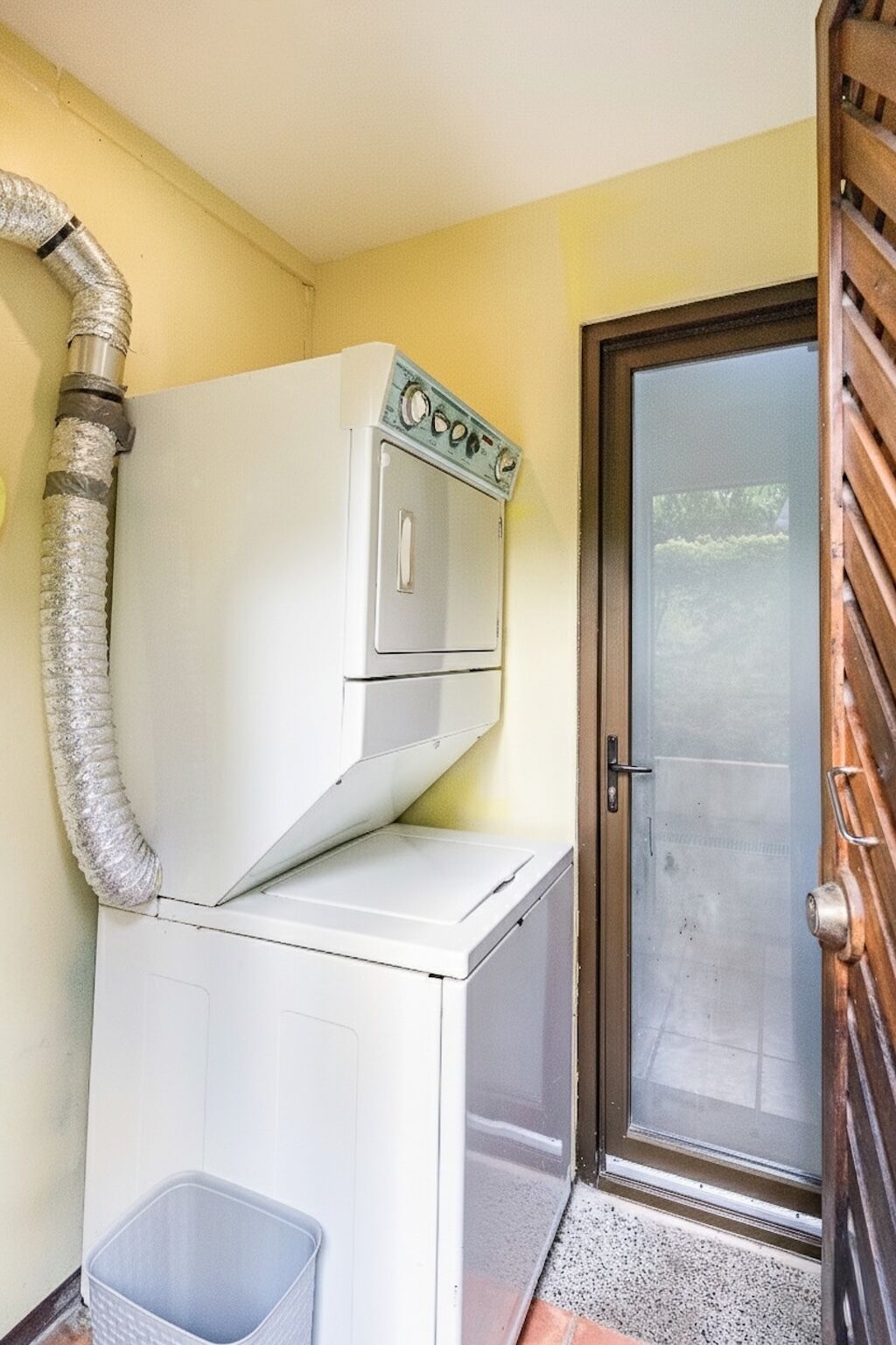 A laundry room with a washing machine and dryer, a vent pipe, a door, and a small trash bin.