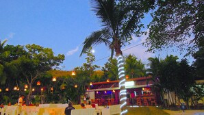 An outdoor seating area with white tables and chairs, a palm tree, and a building with a stage in the background. - PRIVATE ROOFTOP, HILLTOP PANORAMIC OCEAN-VIEW CONDO. COCO SUNSET HILLS, CR. (playas del coco)