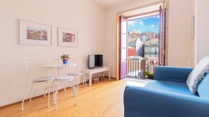 Un salon avec un canapé bleu, des fauteuils blancs, un téléviseur et un balcon offrant une vue sur les immeubles.