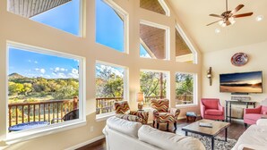 A spacious living room with large windows, a ceiling fan, and a view of a natural landscape. - LAURA’S LODGE – BEAUTIFUL RIVER, KAYAKS, TUBES, HOT TUB - WELCOME! (Wimberley)