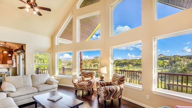 A spacious living room with a high ceiling, large windows, and a view of a lush landscape. - LAURA’S LODGE – BEAUTIFUL RIVER, KAYAKS, TUBES, HOT TUB - WELCOME! (Wimberley)