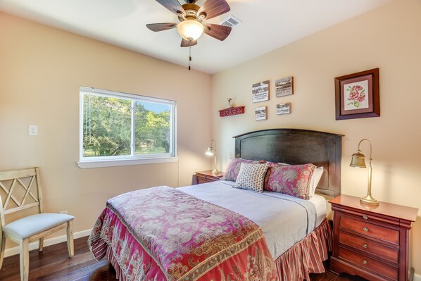 A bedroom with a bed, a nightstand, a chair, a window with a view of trees, and a ceiling fan.