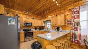 A kitchen with wooden cabinets, a stainless steel refrigerator, a microwave, and a coffee maker. There are wooden stools around the island. - Smoky Mountain Lodge | 6 King Suites, Hot Tub, Views, Game Room, flat entry (Sevierville)