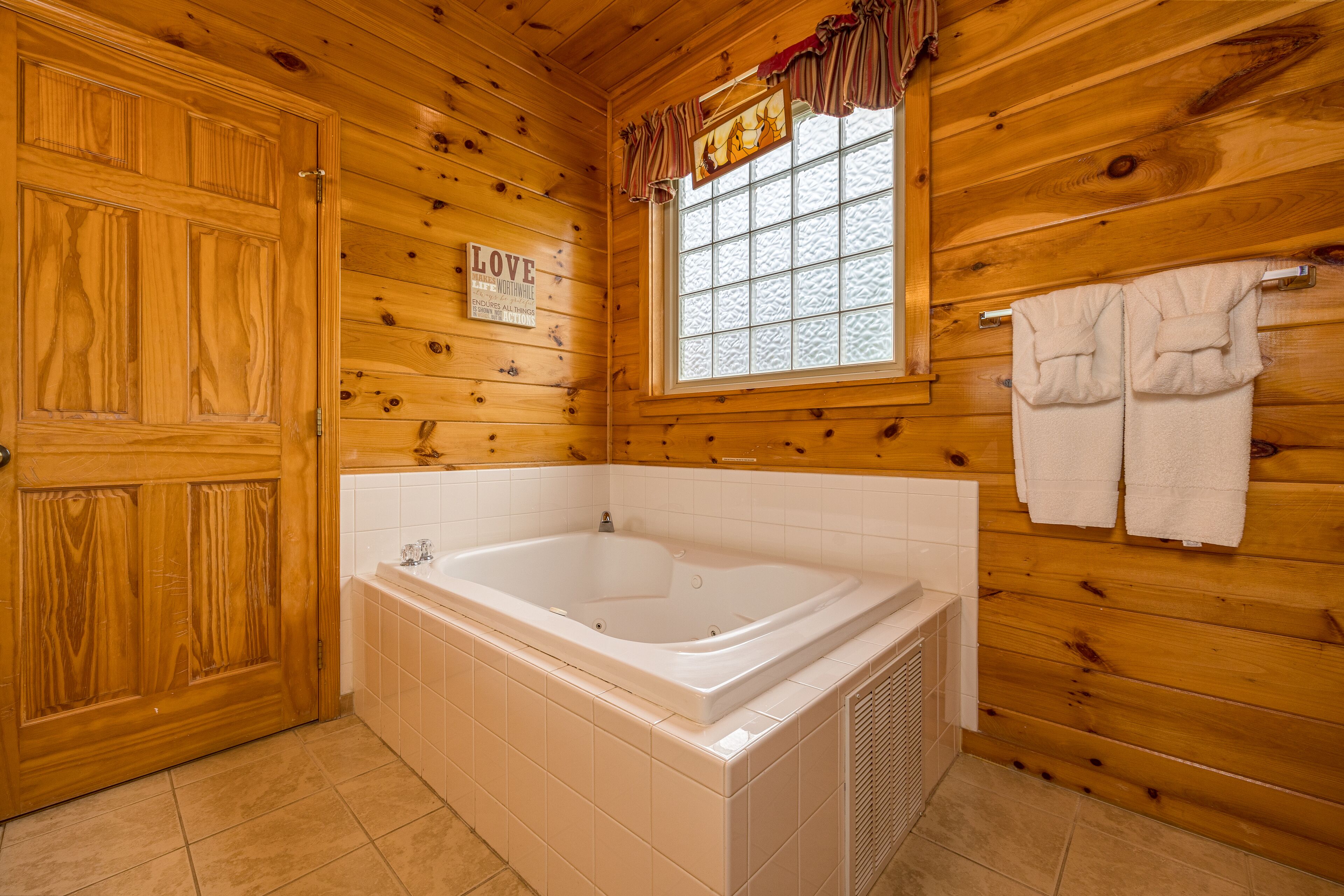 A wooden cabin bathroom with a large bathtub, a window with curtains, and a sign that says "LOVE".