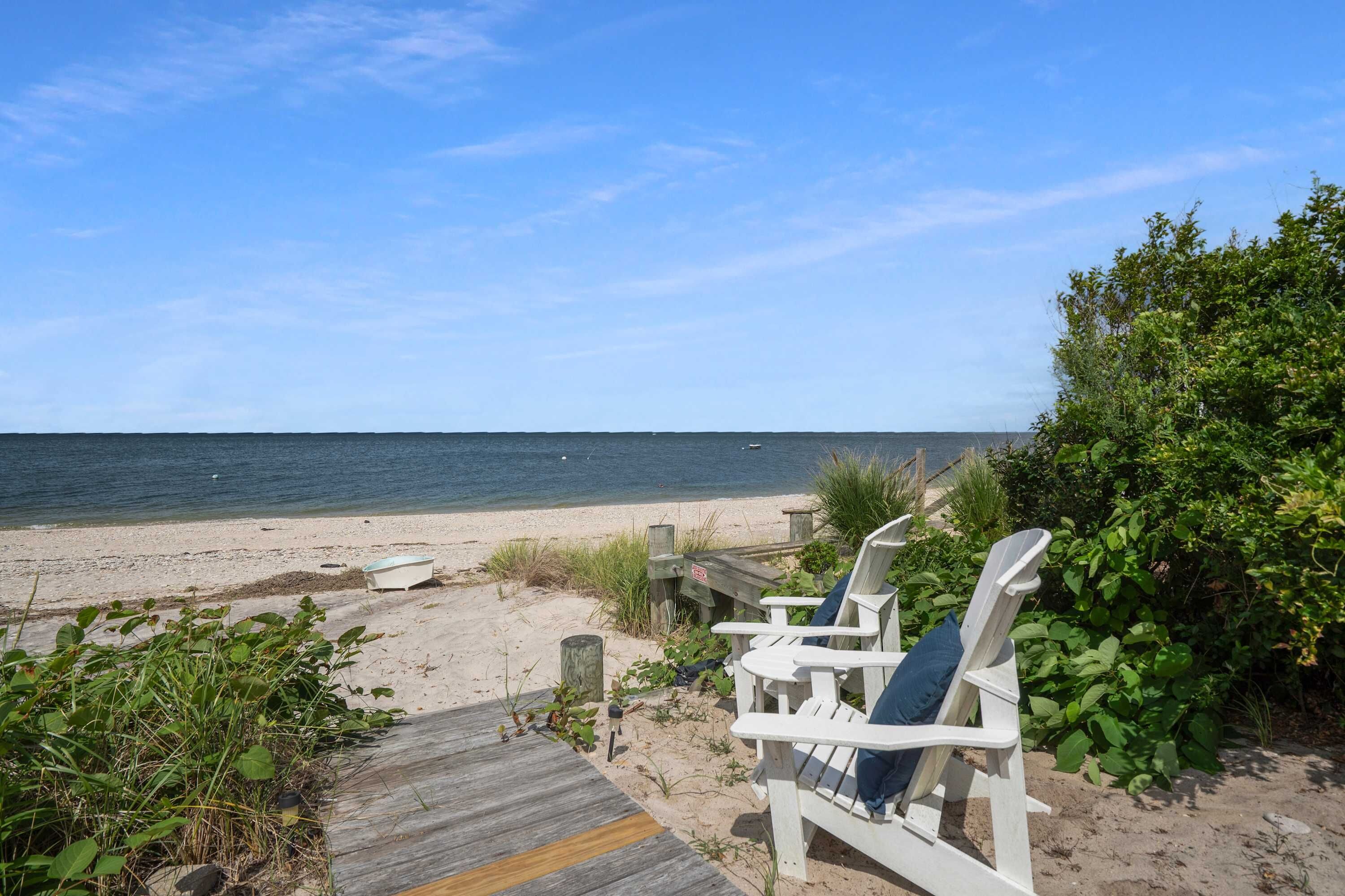A beach with a wooden walkway leading to white Adirondack chairs.