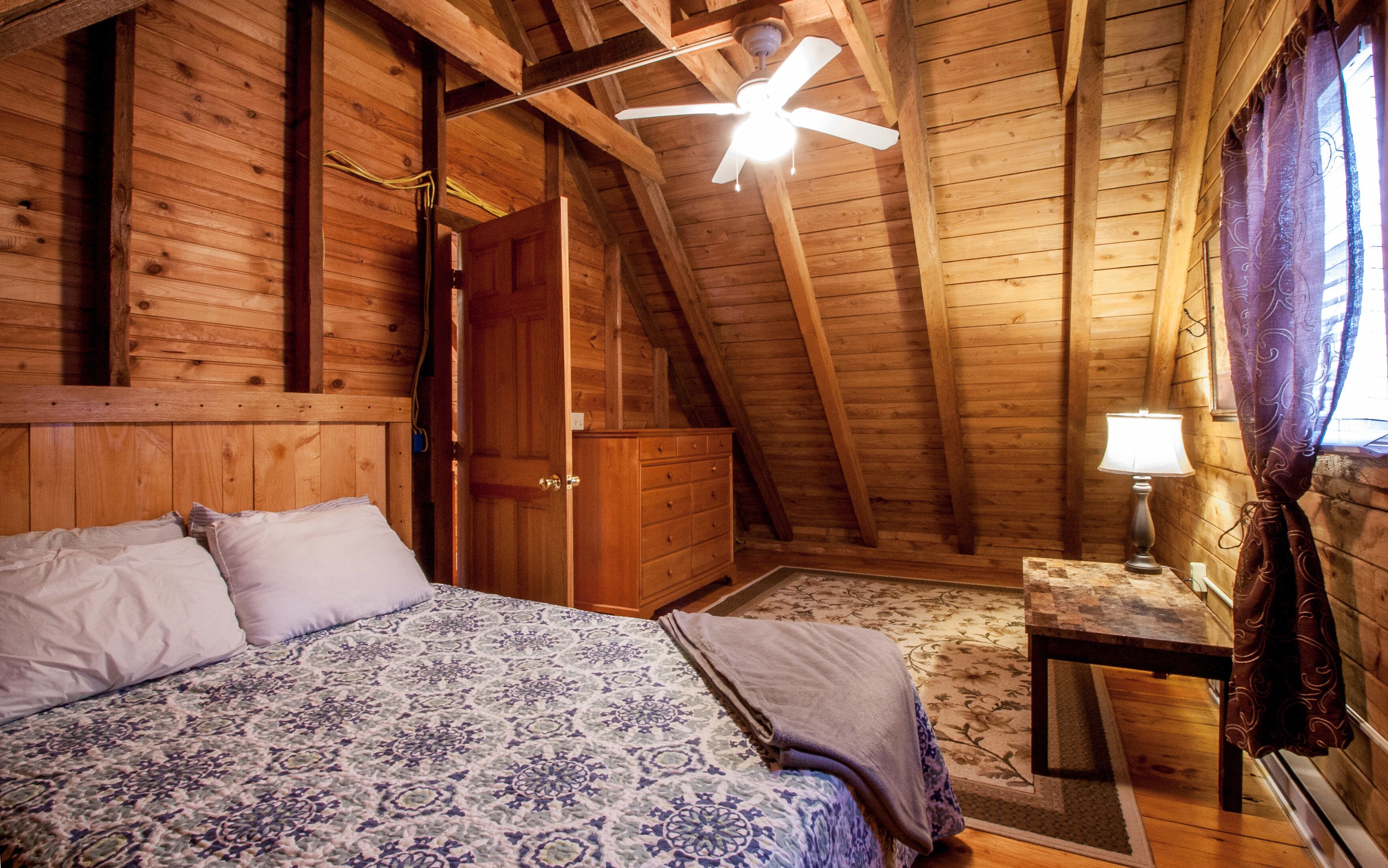 A cozy attic bedroom with a wooden ceiling, a bed with patterned bedding, a dresser, and a small table with a lamp.