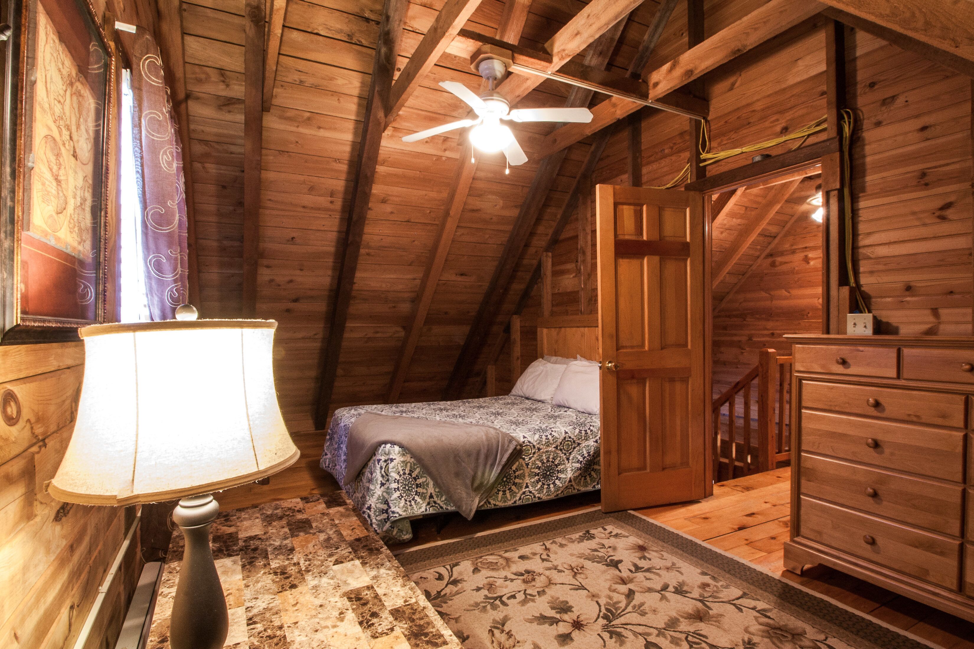 Une chambre mansardée chaleureuse, avec un lit, une commode, une lampe et un ventilateur de plafond.