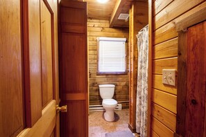 A wooden-paneled bathroom with a toilet, a window with blinds, and a shower curtain.