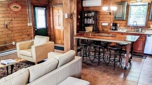 Interior acogedor de una cabaña de troncos con sala de estar que incluye un sofá y sillones, un área de cocina con barra, y una mesa de comedor.
