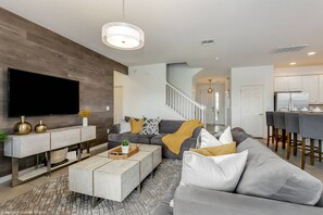 A modern living room with a flat-screen TV, grey sofas, a wooden coffee table, and a staircase leading to a kitchen.