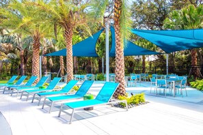 A poolside area with sun loungers and tables, surrounded by palm trees and shaded by blue canopies.