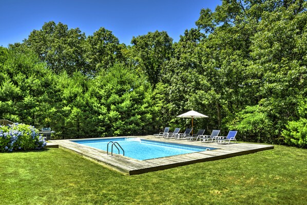 Una piscina rectangular con cubierta de madera, rodeada de césped y árboles.