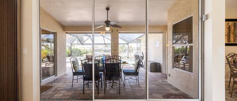 A patio with a table and chairs, a ceiling fan, and a view of a pool and greenery.