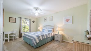 Een slaapkamer met een bed, bureau, stoel, plafondventilator en raam met gordijnen.