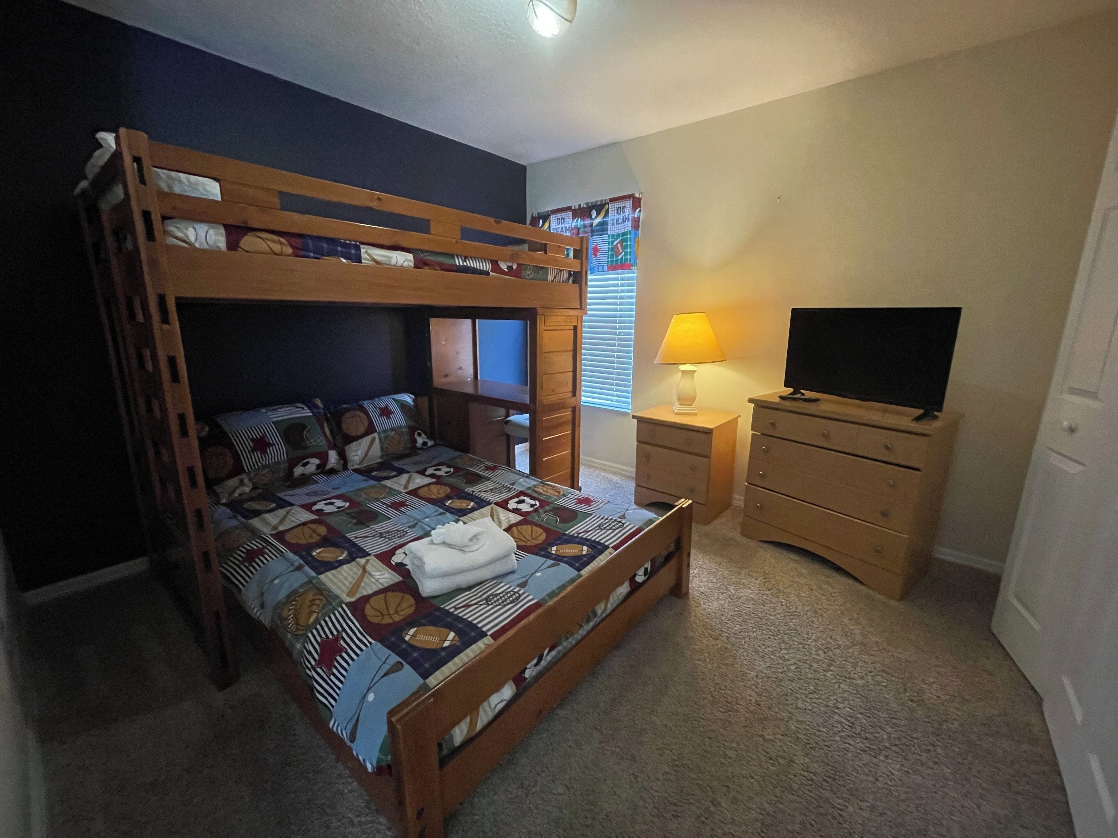A bunk bed with a patterned bedspread, a bedside table with a lamp, a dresser, and a television.