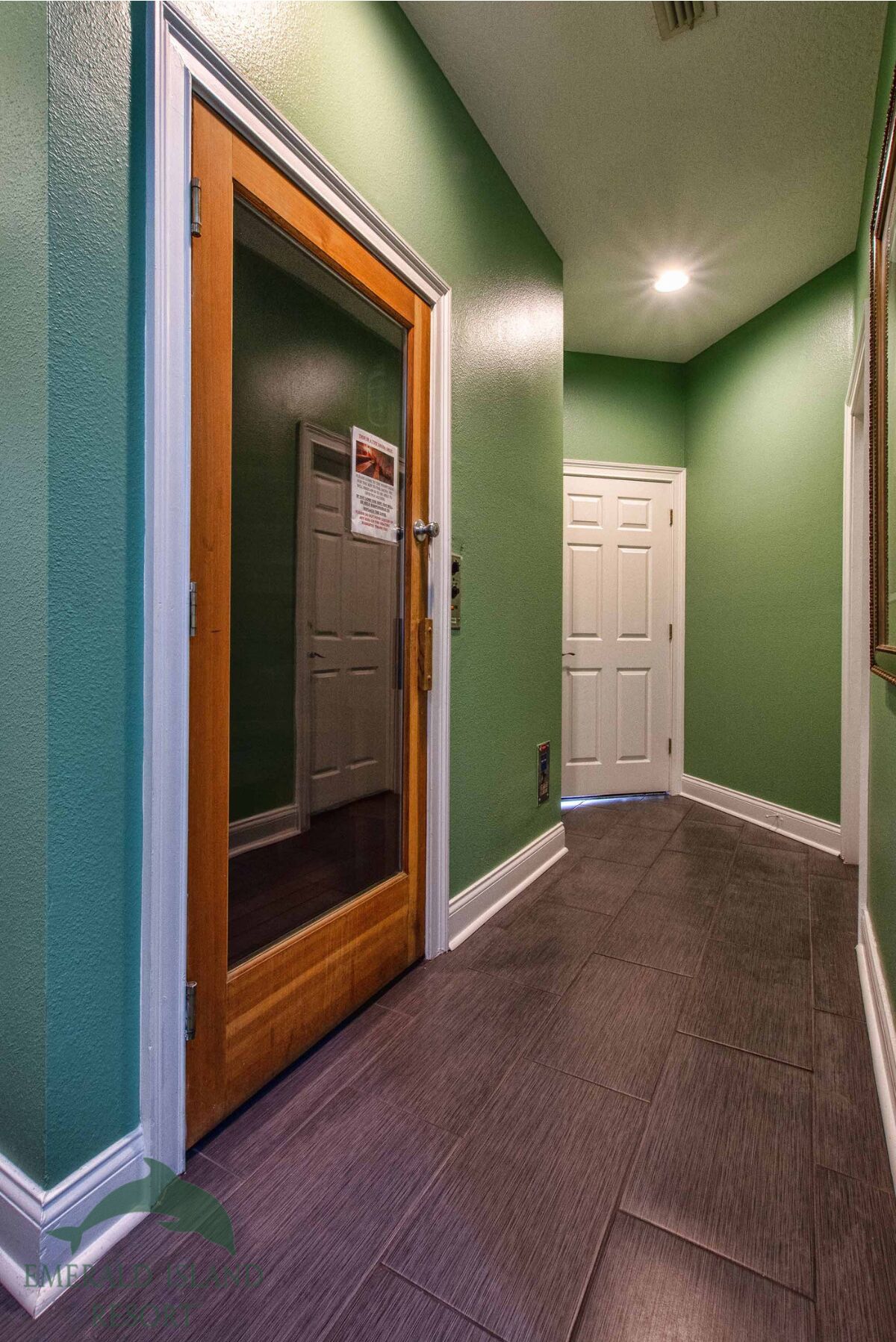 Un couloir aux murs verts, une porte en bois avec un panneau de verre, et une porte blanche au fond.