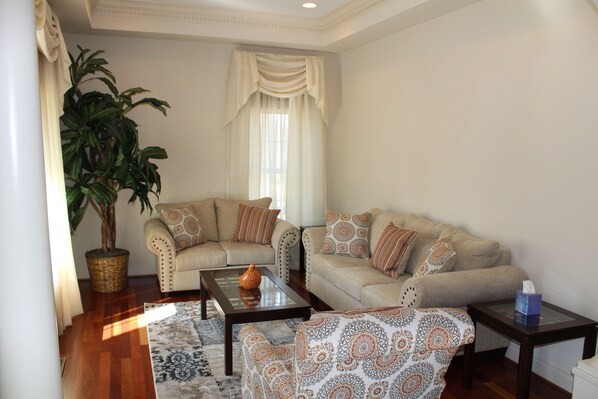 A living room with two sofas, a coffee table, a patterned ottoman, a potted plant, and a window with curtains.