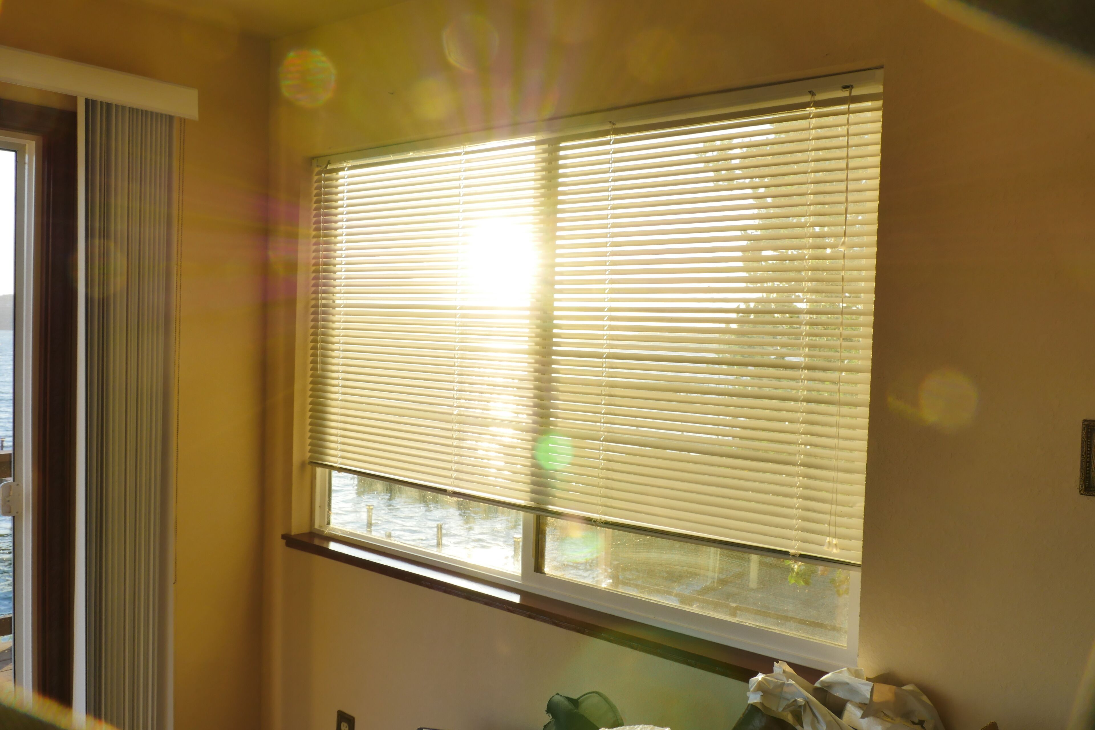 A room with a large window with blinds partially open, a door with a view of the outside, and a small table with a green object on it.