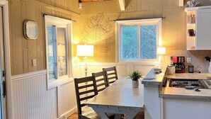Un coin repas chaleureux avec une table et des chaises en bois, une cuisine équipée d’une cuisinière et une fenêtre donnant sur les arbres.