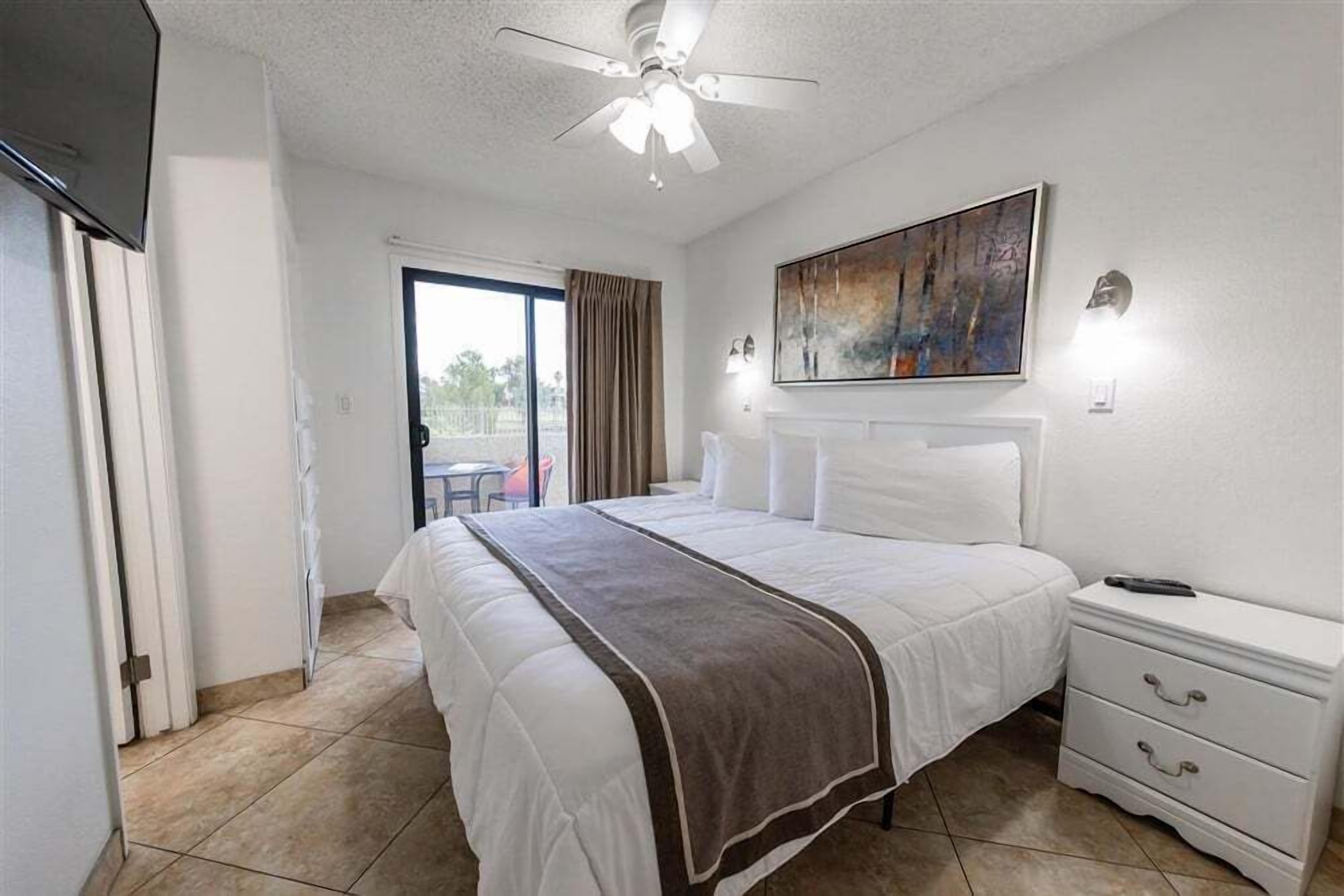 A hotel room with a bed, a nightstand, a ceiling fan, a painting on the wall, and a view through the door.