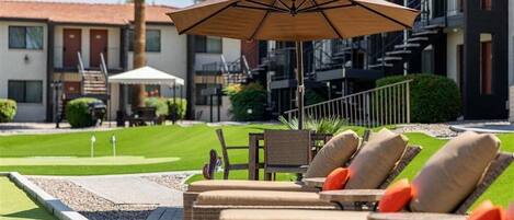 A poolside area with lounge chairs and an umbrella, a well-maintained lawn, and a building with balconies in the background.