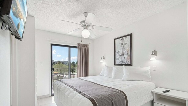 A bedroom with a bed, a ceiling fan, a TV, a painting, and a balcony with a view.