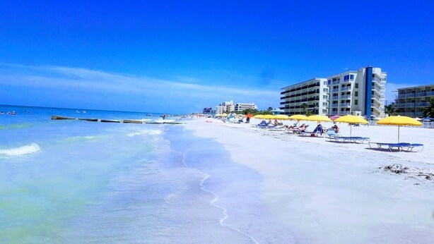A beach with white sand, clear blue water, and a row of yellow umbrellas. There are buildings in the background.