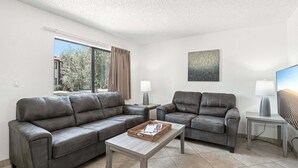 A modern living room with two grey sofas, a coffee table, two side tables with lamps, a painting on the wall, and a window with curtains.