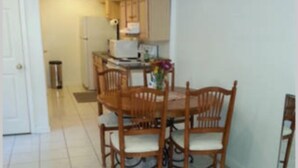 A dining area with a wooden table and chairs, a kitchen in the background, and a tiled floor.