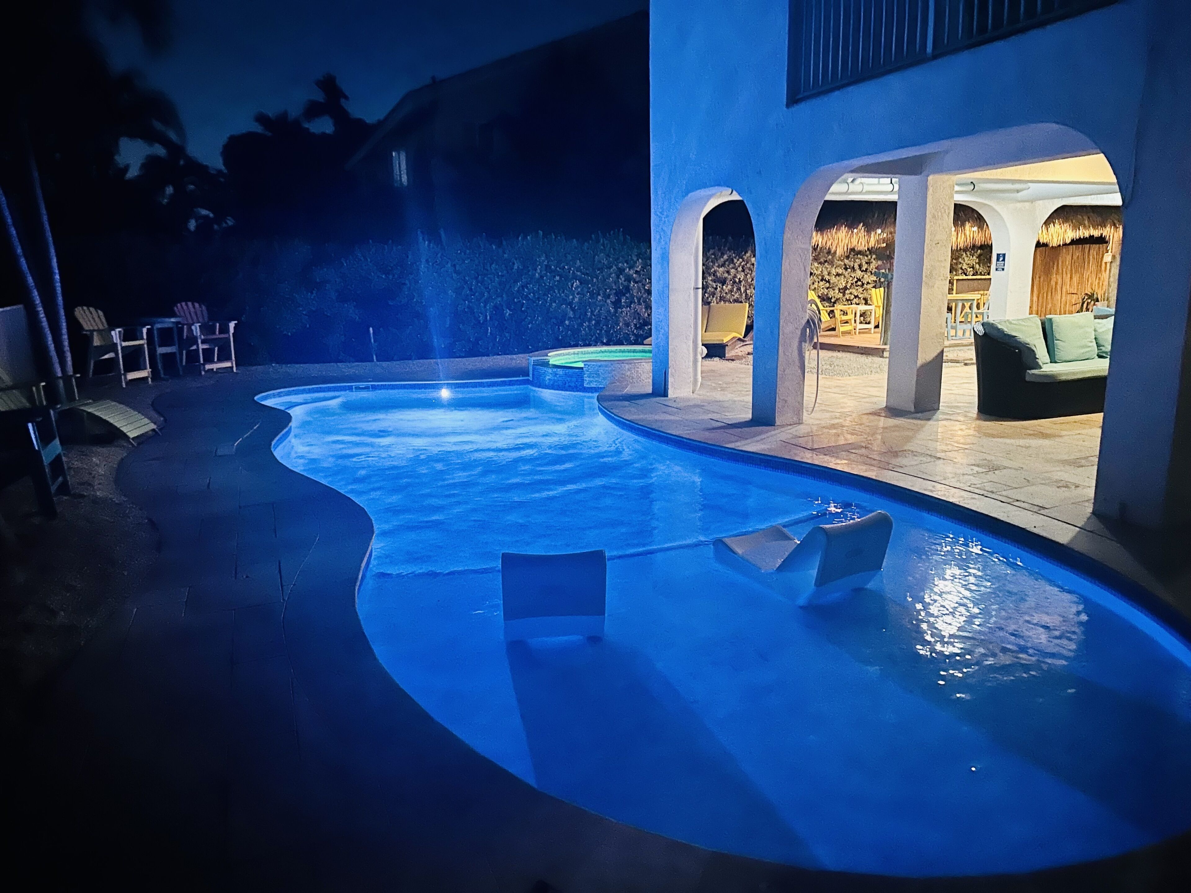 A swimming pool with a curved design, surrounded by outdoor seating and a covered area with a thatched roof.