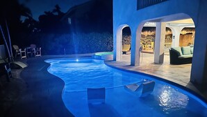 Une piscine au design incurvé, entourée d’espaces de détente en plein air et d’une zone abritée avec un toit de chaume.