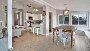 Cozinha moderna com armários brancos, uma ilha central e banquetas. Há uma sala de jantar com mesa de madeira e cadeiras brancas.